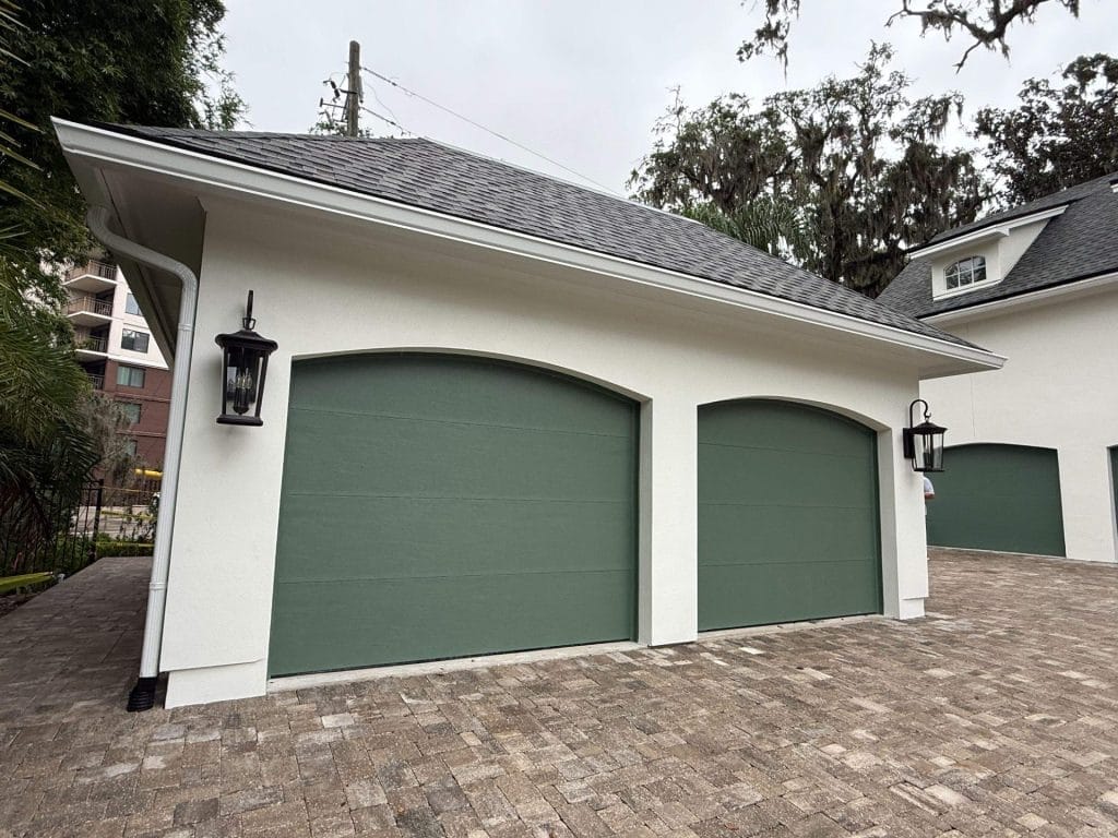 Custom two-car garage addition with arched green doors, white stucco siding, and a paver driveway by The Dantzler Group in Jacksonville, FL.